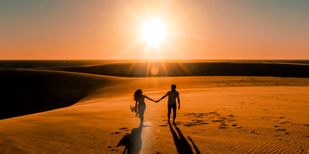Vandressa e Júnior caminham de mãos dadas, em silhueta, com o sol entre as dunas.