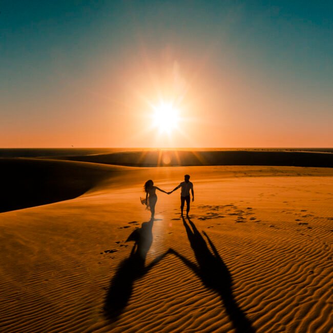 Vandressa e Júnior caminham de mãos dadas, em silhueta, com o sol entre as dunas.