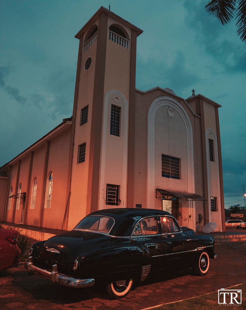 Um carro preto antigo fica diante de uma igreja com torre.