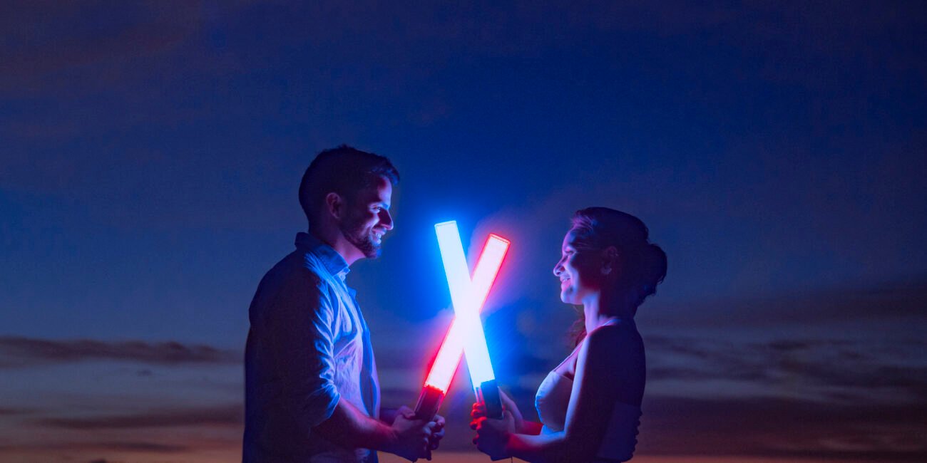 Ana e Arthur com sabres de luz azul e vermelho cruzados na praia à noite