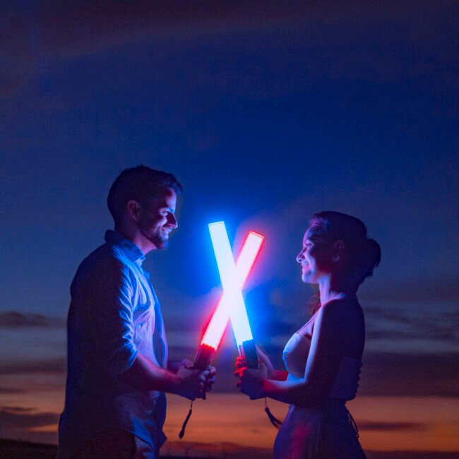 Ana e Arthur com sabres de luz azul e vermelho cruzados na praia à noite