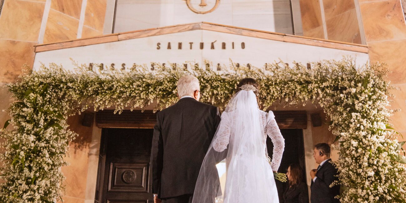 Armeny, de véu e vestido de renda, entra de costas pela porta onde se lê SANTUÁRIO.