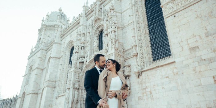 Sara e Daniel se olham junto à fachada, ela de casaco bege e ele de terno preto.