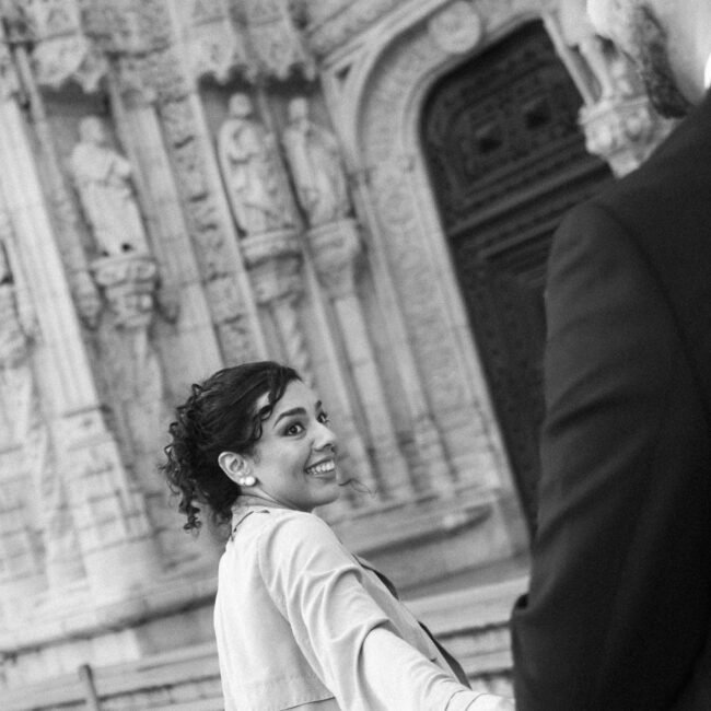 Sara sorri para trás, de casaco bege, segurando a mão de Daniel.