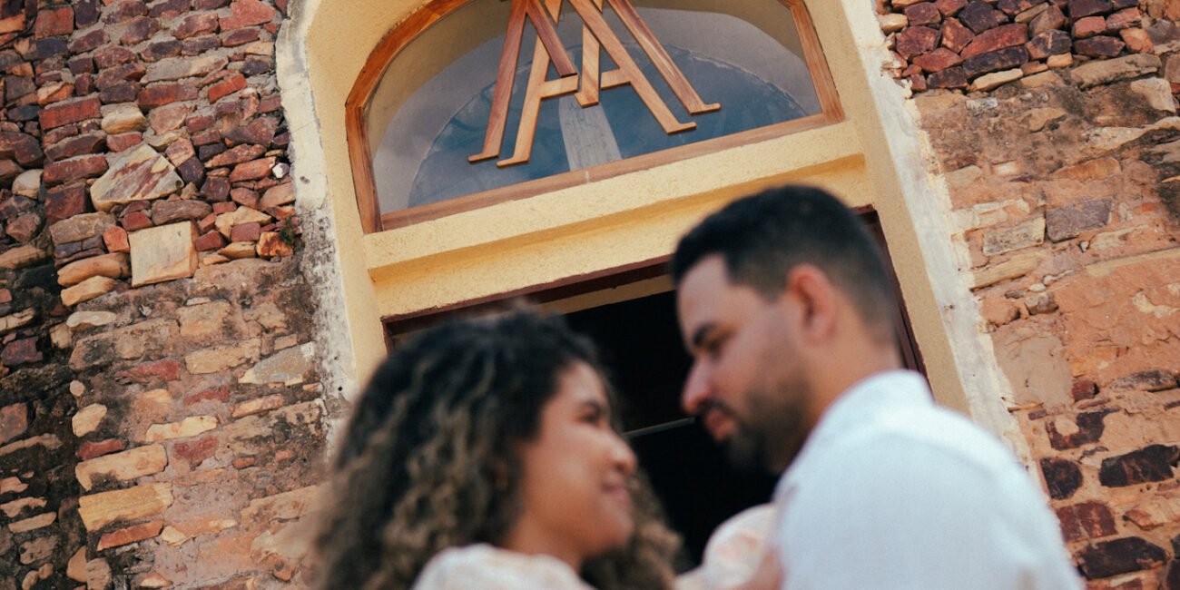 Andressa e Fábio se abraçam na porta, sob a cruz e a janela em arco.