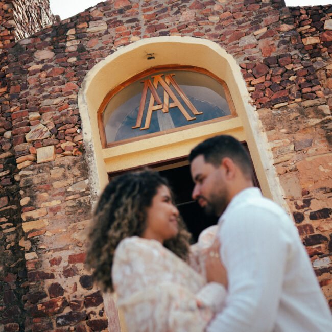 Andressa e Fábio se abraçam na porta, sob a cruz e a janela em arco.