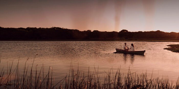 Raquel e Thiago estão num barco no lago, pequenos diante da mata.